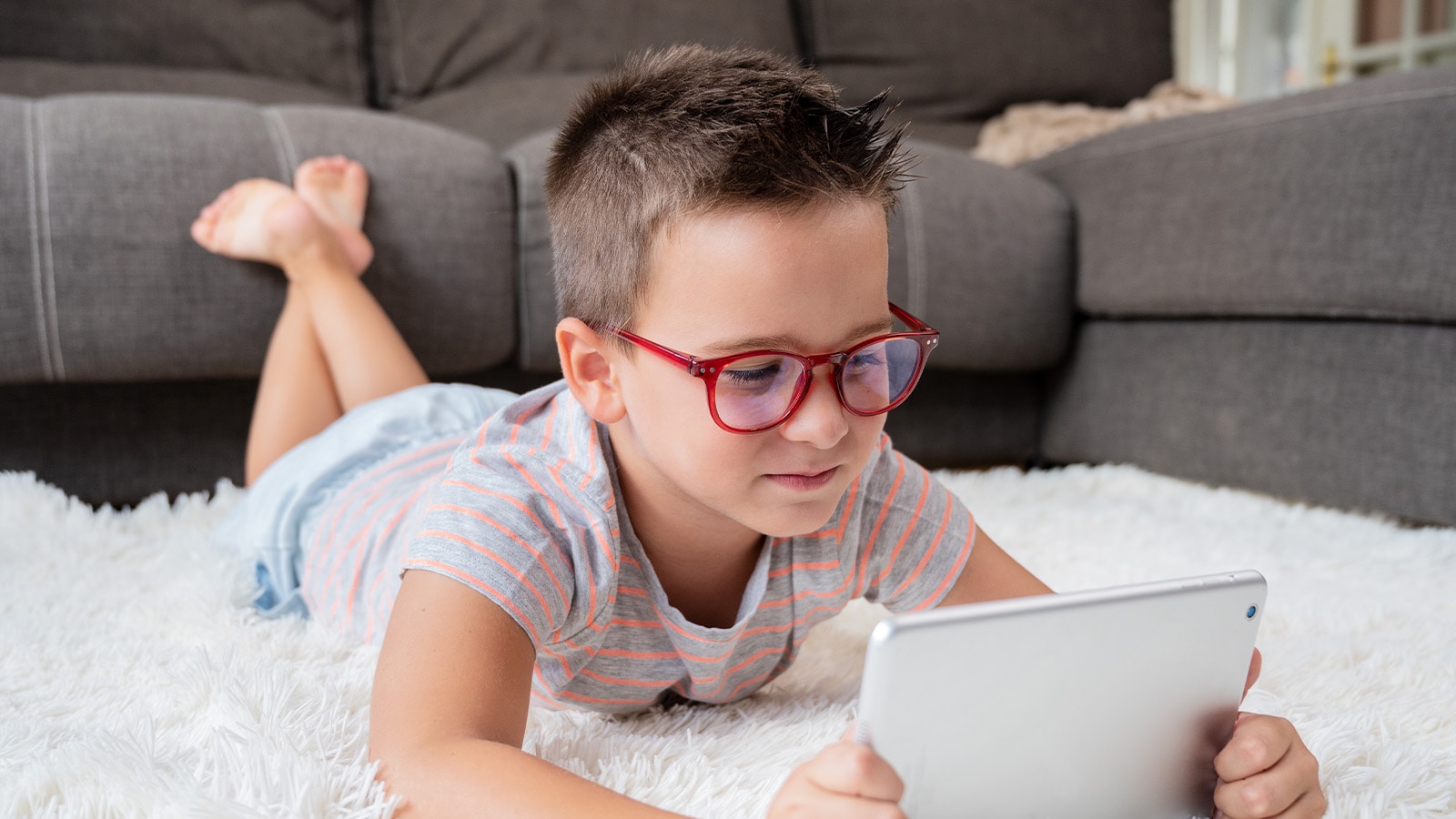 Young boy in red glasses reading a tablet, showing a child needs glasses for clear focus.