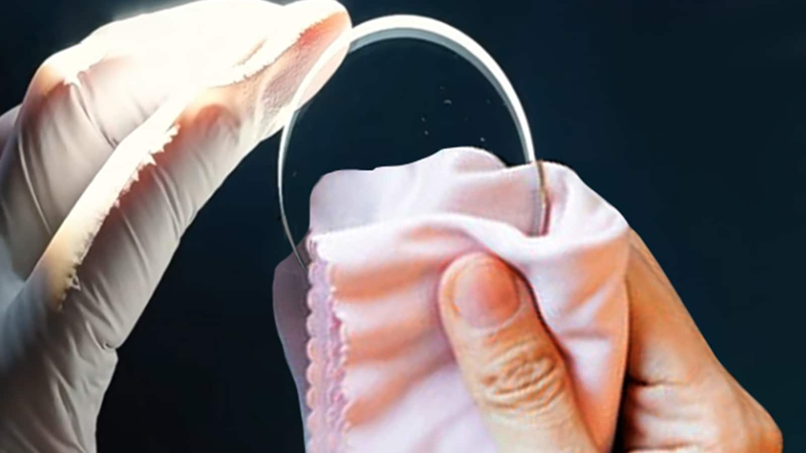 Hands in gloves cleaning a detached glasses lens with a soft cloth before reinsertion.