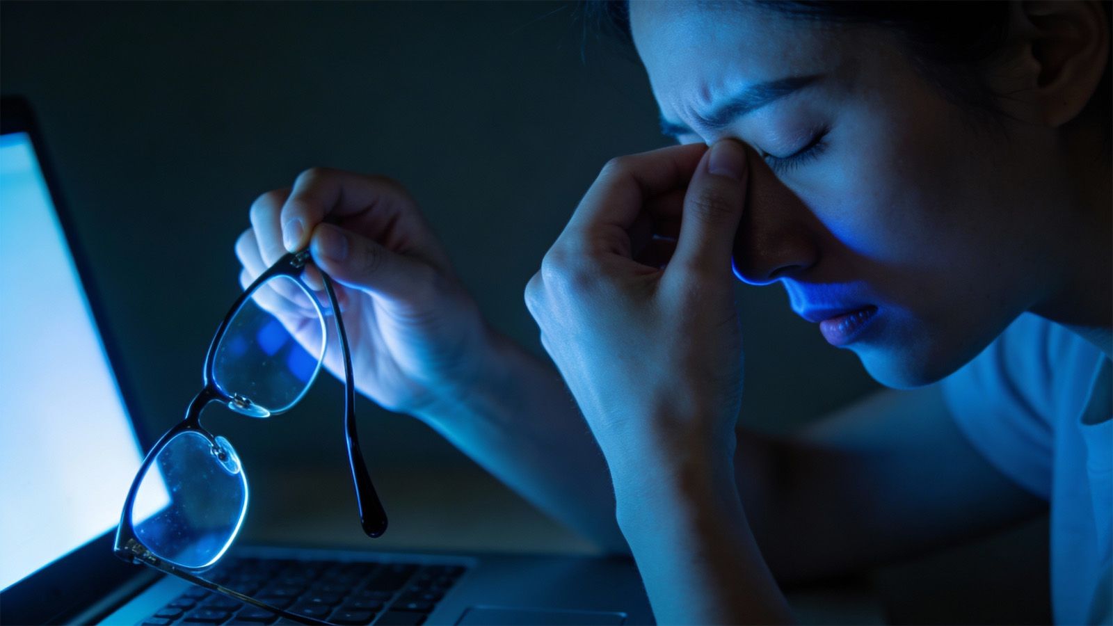Woman at laptop rubbing tired eyes holding glasses indicating digital eye strain from prolonged screen time and need for updated lenses.