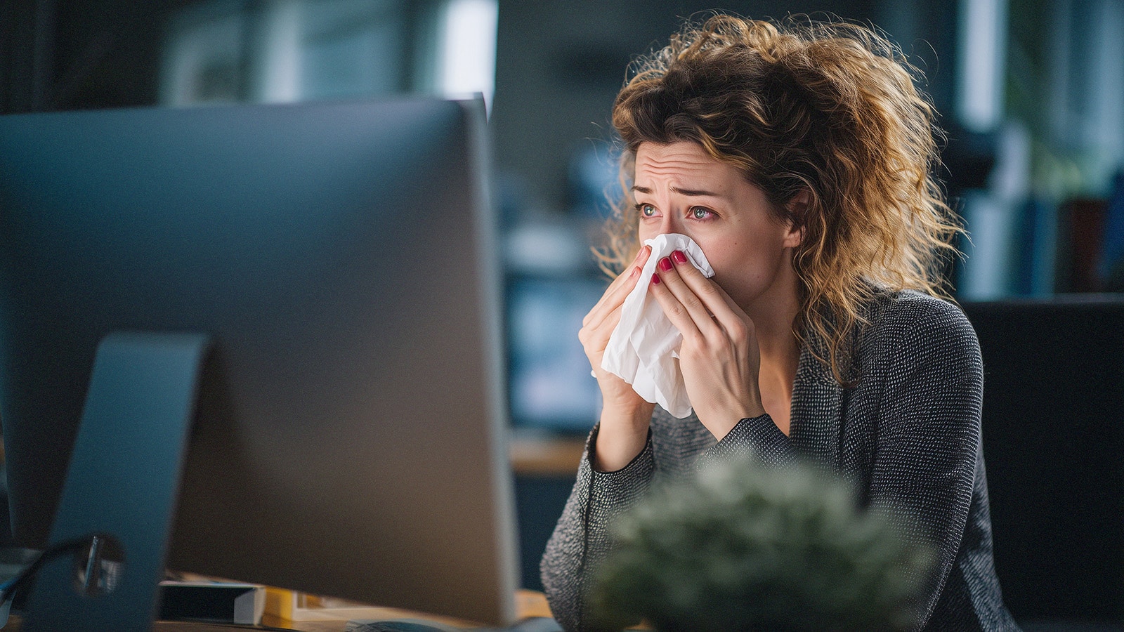 Woman rubbing eyes and using tissue at computer, showing discomfort and irritation from dry eye syndrome.