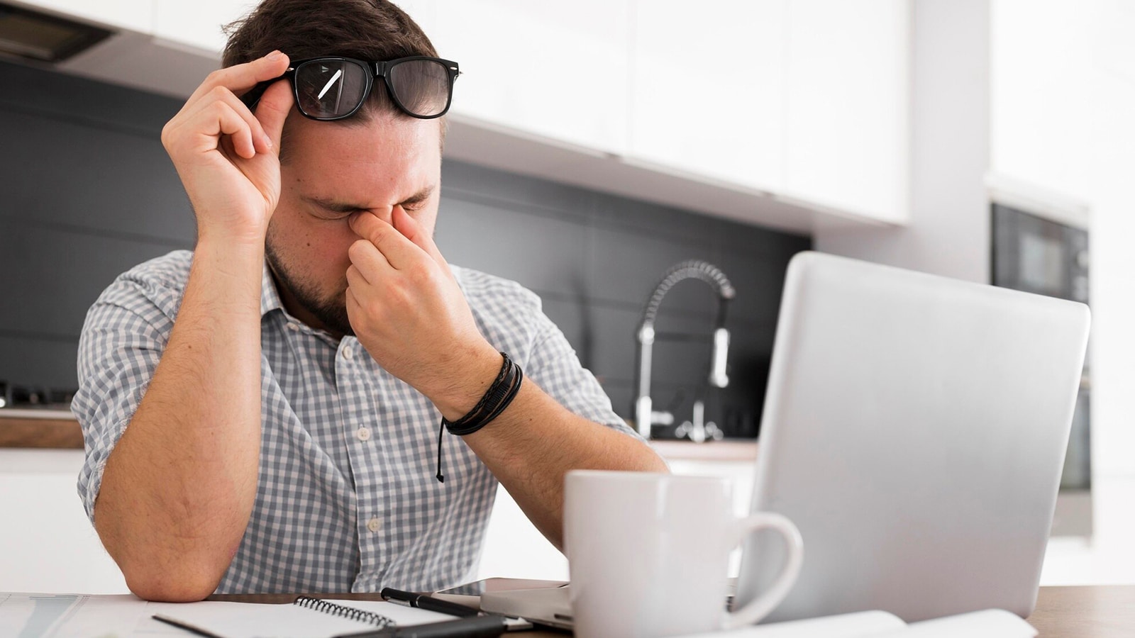 A man with glasses pushed up on his forehead, rubbing his eyes due to digital eye strain while sitting at a desk with a laptop and a mug.