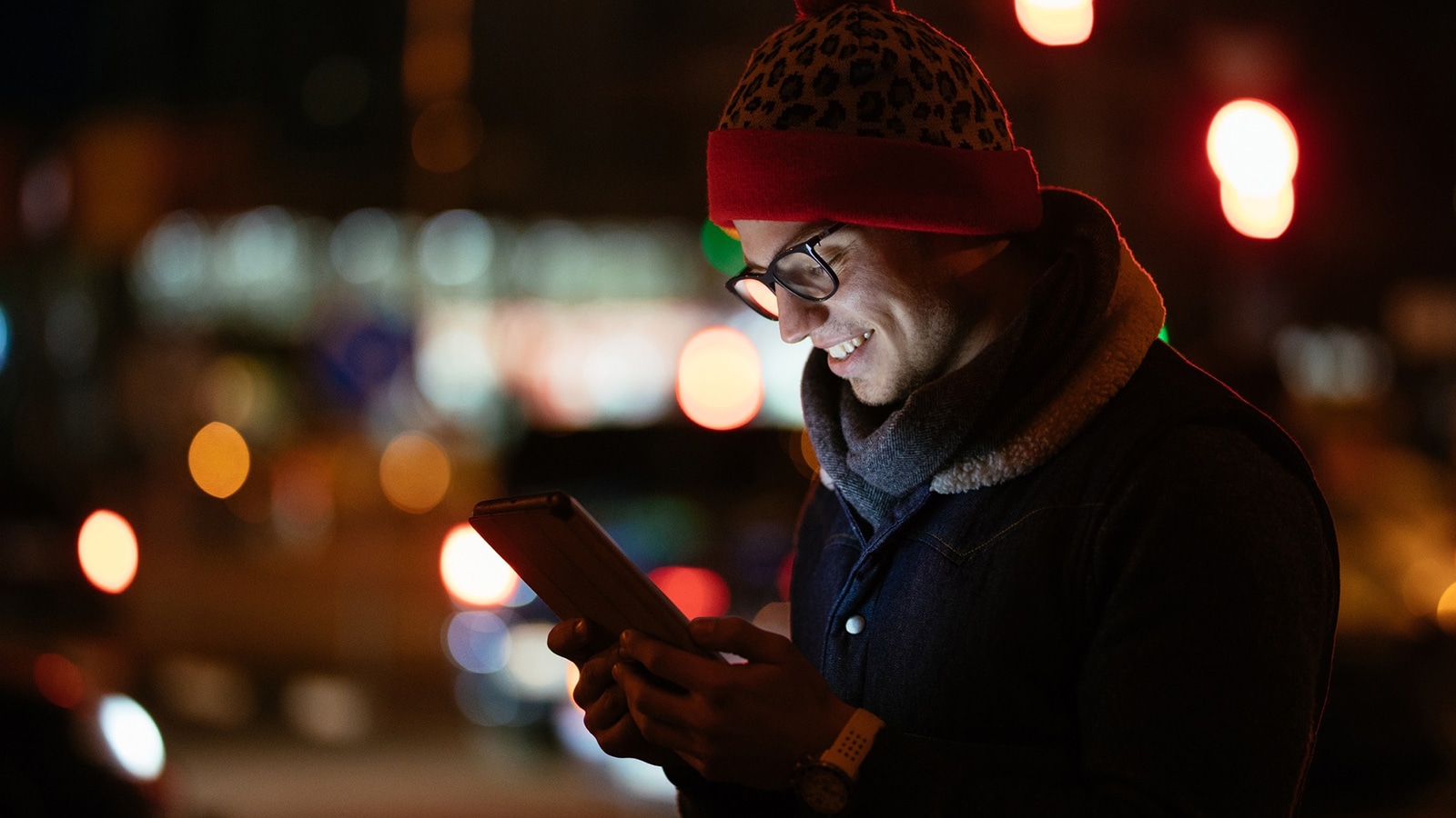 A man wearing glasses and a hat, smiling while looking at a tablet in a dimly lit urban environment at night.