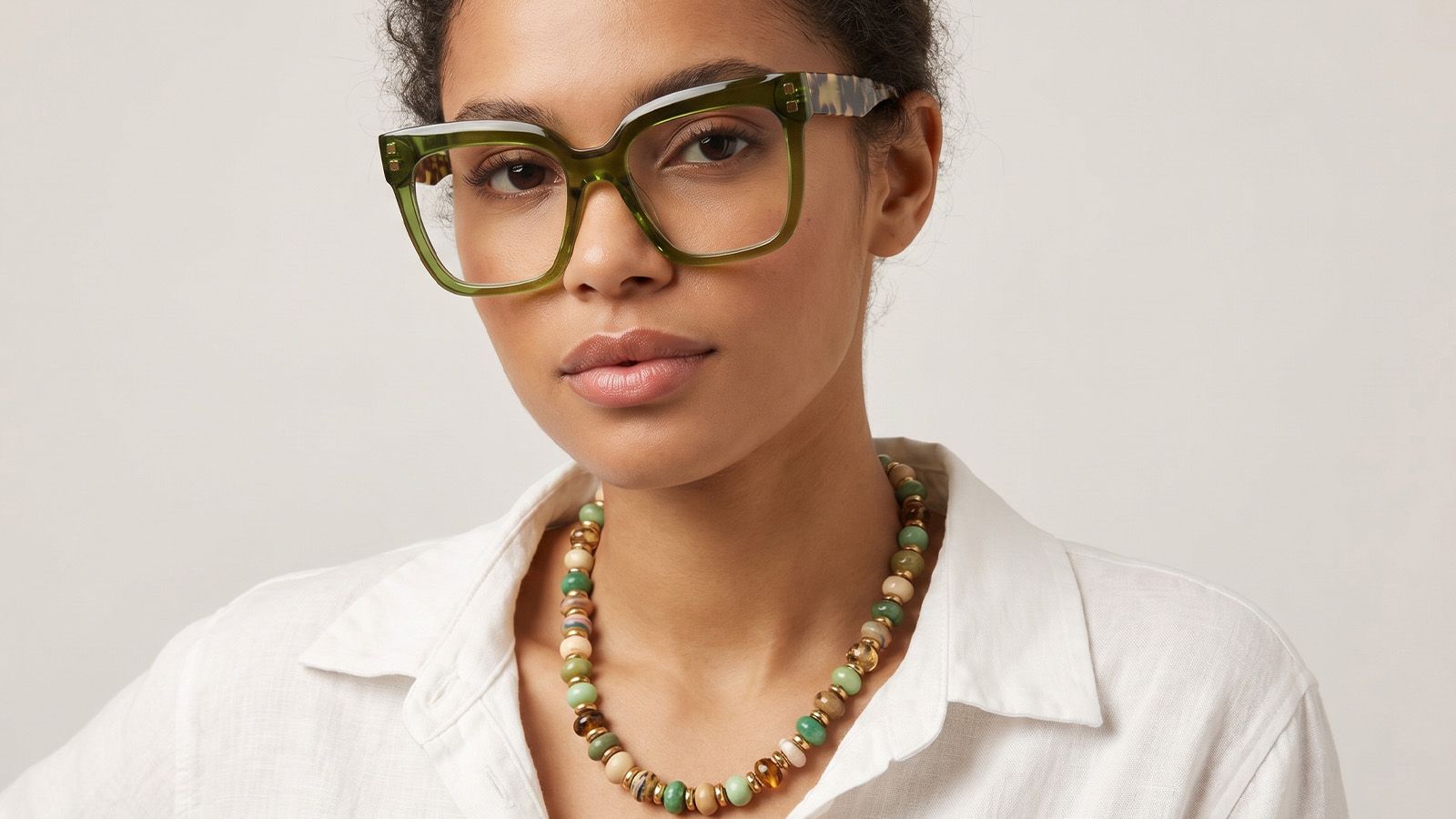 Close-up of a model wearing bold neon green transparent acetate eyeglasses and a minimalist white linen shirt. Layered colorful beaded necklaces complement the eyewear, highlighting HD detail on professional glasses and modern accessory trends.