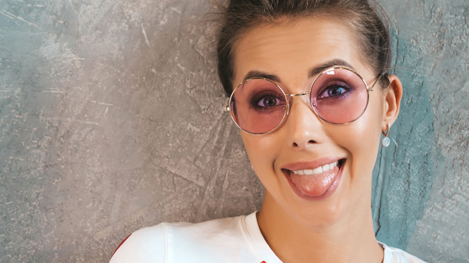 Smiling woman in round pink-tinted glasses sticking out tongue, highlighting fun eye makeup balance with bold frames.