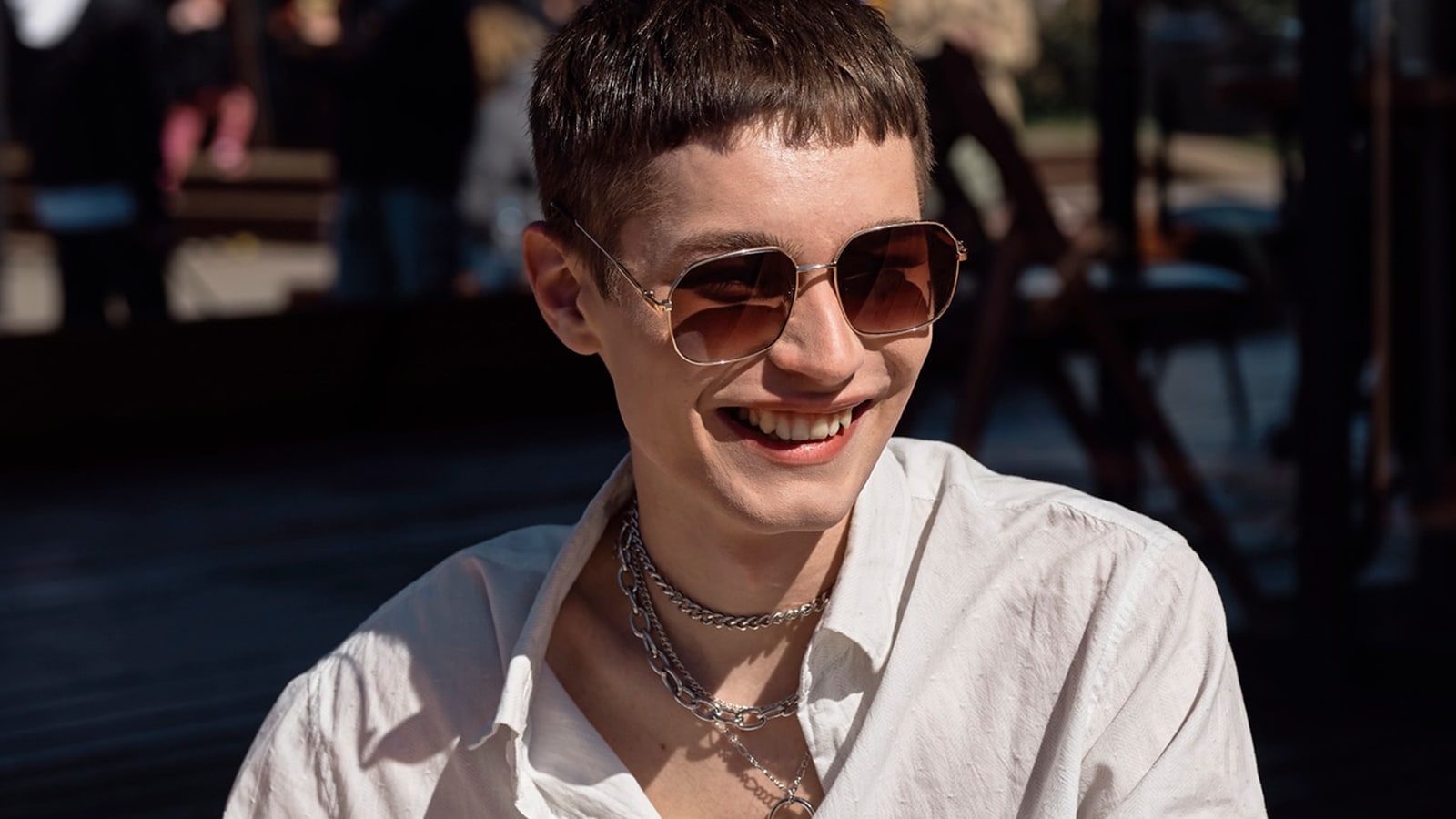A smiling person wearing oversized titanium sunglasses and a chain necklace.