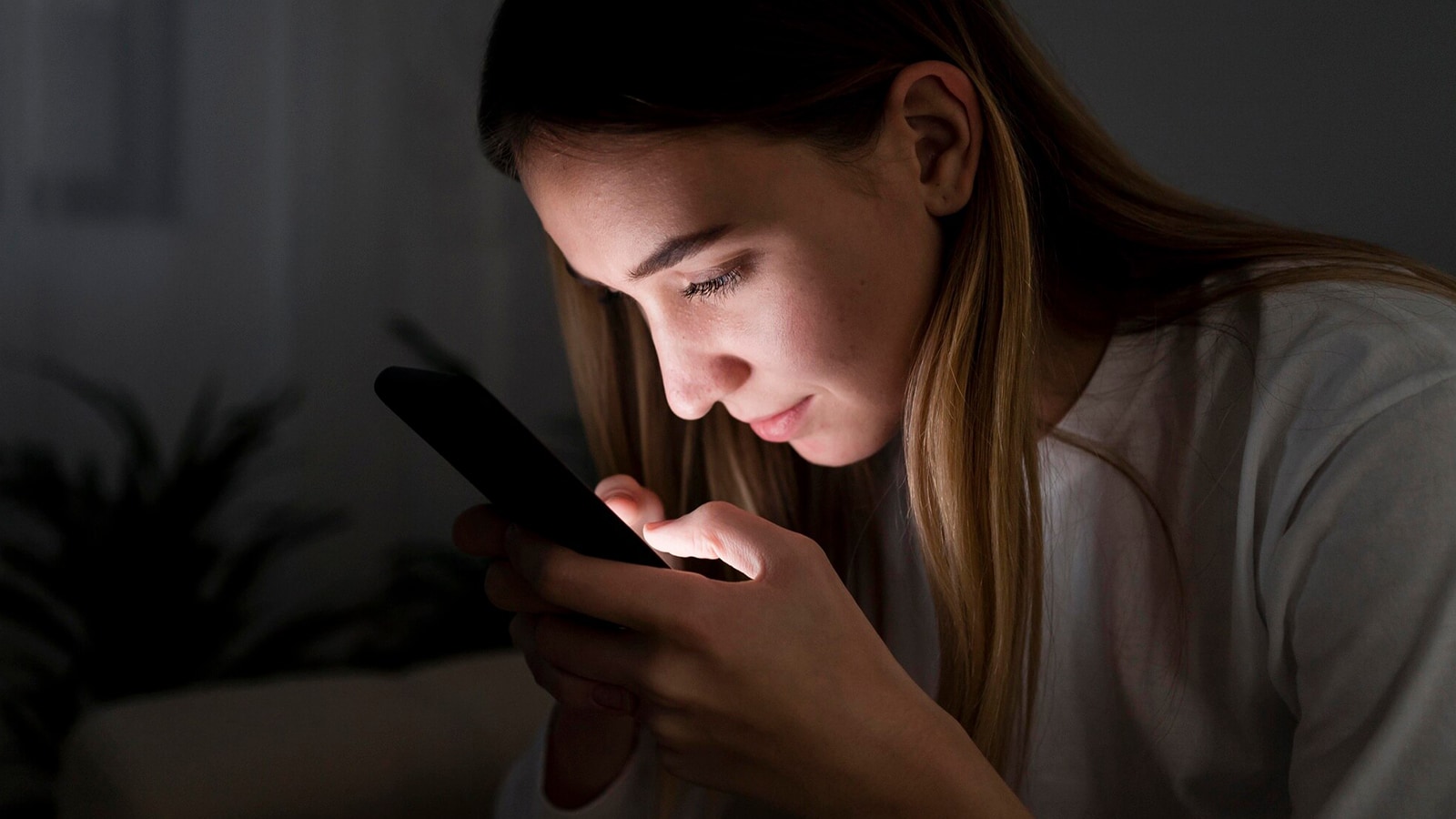 A woman holding her phone in a dark room, with the screen light on her face, appearing focused.