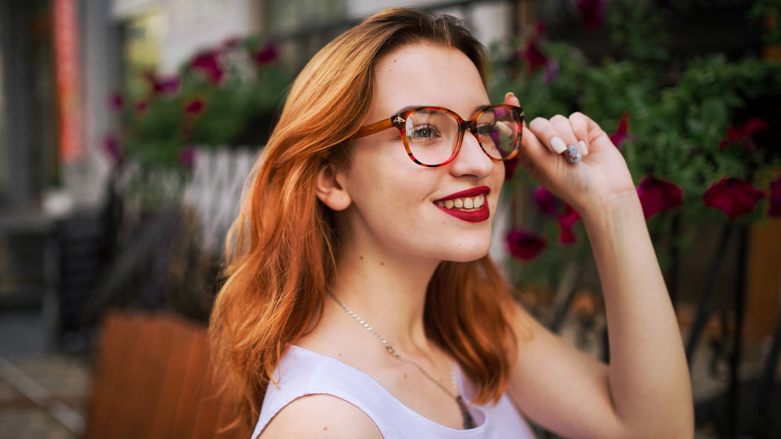 A stylish woman with red hair smiles while adjusting her tortoise-shell glasses, demonstrating a comfortable fit for a high bridge nose.