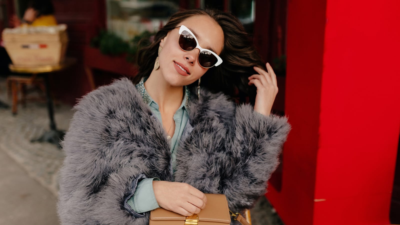 A woman in a faux fur coat and white oversized glasses sitting at a cafe table.