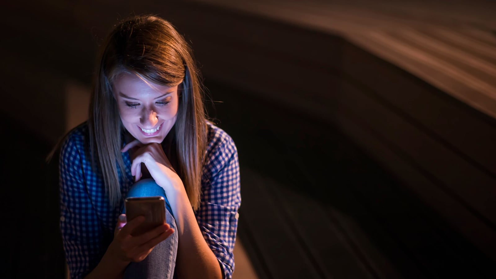 A young woman smiling while looking at her phone in a dimly lit room, with the phone's light illuminating her face.