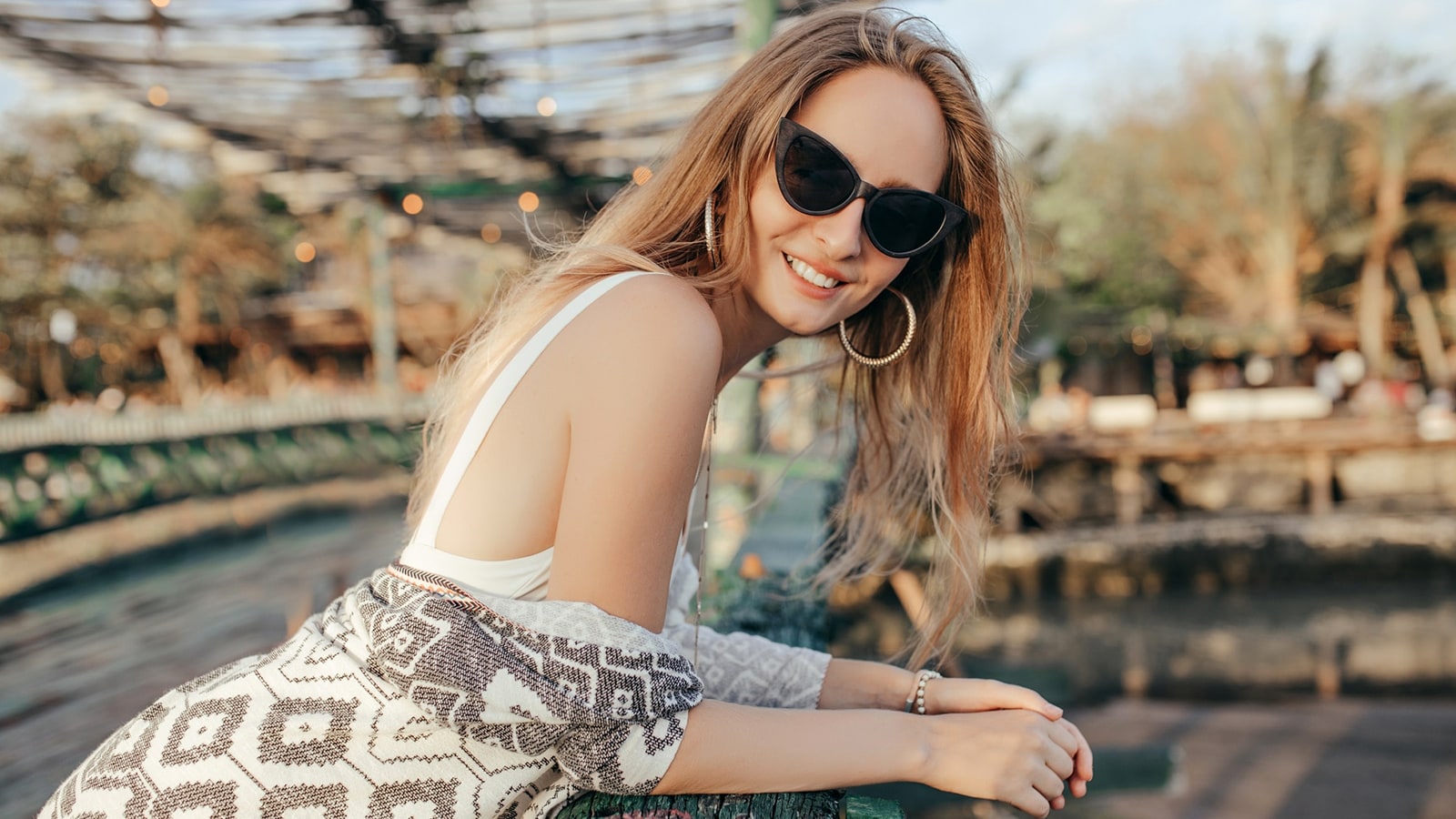A smiling woman with blonde hair and oversized glasses resting on a railing outdoors.