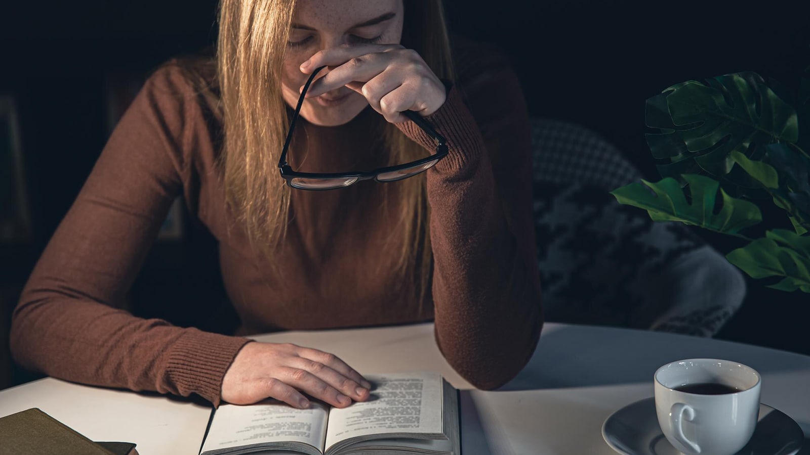 A woman reading a book in a dark room, holding her glasses and rubbing her eyes, showing signs of eye strain.