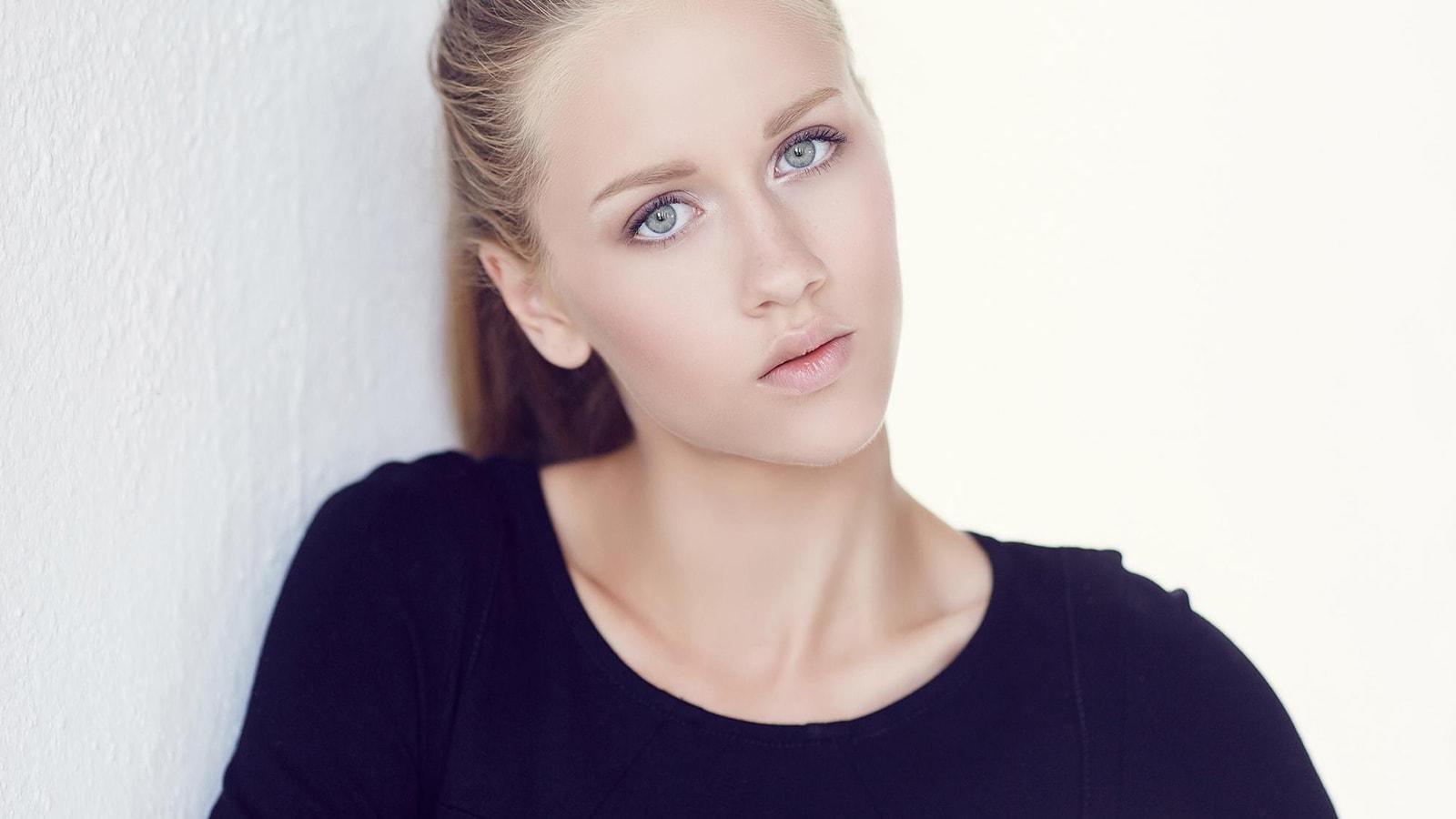 A woman with light, possibly grayish-blue eyes, wearing a black top, leaning against a white wall with a soft expression.