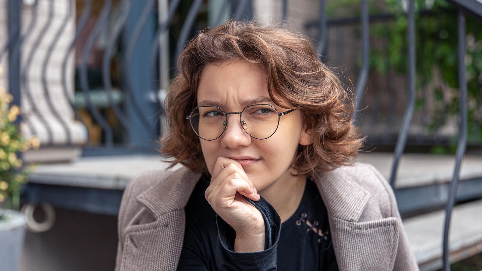 A young woman with brown, wavy hair wearing round glasses, resting her chin on her hand in a thoughtful pose.
